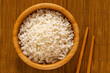 Cooked white rice in a dark wood bowl next to chopsticks isolated on bamboo matt from above.