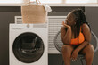 © gstockstudio - Bored curvy African woman waiting for clean clothes while sitting in chair near the washing machine in laundry