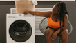 © gstockstudio - Young African woman select settings for laundry while sitting in comfortable chair near the washing machine