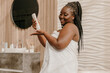 © gstockstudio - Happy plus size African woman covered in towel applying cosmetic cream on hand in bathroom