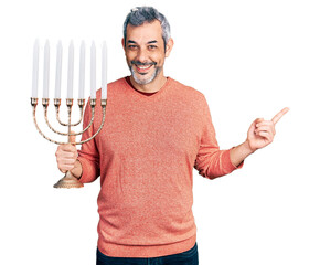 Canvas Print - Middle age hispanic with grey hair holding menorah hanukkah jewish candle smiling happy pointing with hand and finger to the side