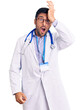 Young hispanic man wearing doctor uniform and stethoscope surprised with hand on head for mistake, remember error. forgot, bad memory concept.