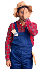 Poster - Handsome latin american young man weaing handyman uniform covering one eye with hand, confident smile on face and surprise emotion.