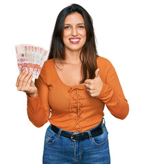 Wall Mural - Beautiful hispanic woman holding 10 colombian pesos banknotes smiling happy and positive, thumb up doing excellent and approval sign