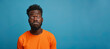 A man with dreadlocks and an orange shirt is staring at the camera. Portrait of a confused puzzled minded African American man in orange top isolated on blue background, copy space.