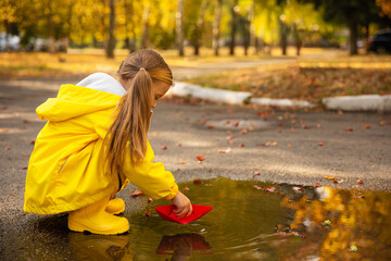 Wall Mural - Happy child girl in a yellow jacket and yellow rubber boots has fun with paper boat in a puddle in autumn on nature. Happy childhood