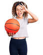 © Krakenimages.com - Beautiful kid girl with curly hair holding basketball ball smiling happy doing ok sign with hand on eye looking through fingers