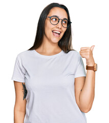 Wall Mural - Young hispanic woman wearing casual white t shirt smiling with happy face looking and pointing to the side with thumb up.