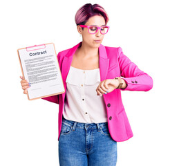 Canvas Print - Young beautiful woman with pink hair holding clipboard with contract document checking the time on wrist watch, relaxed and confident