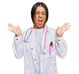 Canvas Print - Beautiful hispanic woman wearing doctor uniform and stethoscope celebrating mad and crazy for success with arms raised and closed eyes screaming excited. winner concept