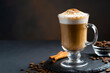 Cappuccino in a clear glass mug, with whipped cream on top, dusted with cinnamon, on a black background.