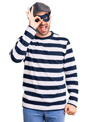 Sticker - Young handsome man wearing burglar mask doing ok gesture with hand smiling, eye looking through fingers with happy face.