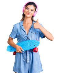 Wall Mural - Young beautiful woman wearing headphones and holding skate smiling happy and positive, thumb up doing excellent and approval sign