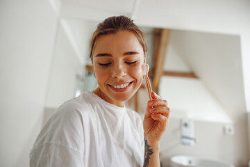 Poster - Beautiful woman use jade roller to massage her face standing in bathroom. Spa self beauty procedure