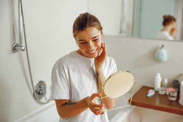 Poster - Smiling young woman looking in small round mirror and checking skin while standing in bathroom