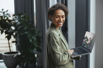 Wall Mural - A woman holding a laptop stands by a window with a houseplant