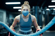 A female athlete is practicing with a battle rope while wearing a protective face mask in a gym