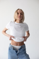 Wall Mural - Beautiful blonde girl in a white T-shirt and blue jeans and sneakers posing on a white background