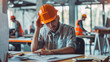 © Kien - A construction worker sitting at his desk, looking frustrated and wearing an orange hard hat on their head
