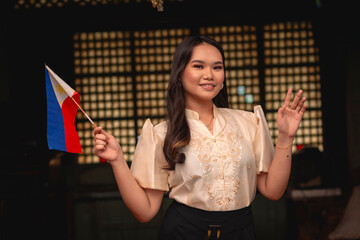 Portrait of a young tour guide holding a Philippine flag, dressed in elegant traditional Filipino attire waving hi. Inside an ancestral house with capiz windows.