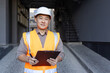 Portrait of a smiling young Asian man standing outside a construction site in a hard hat and vest, holding a folder with documents and smiling, looking at the camera