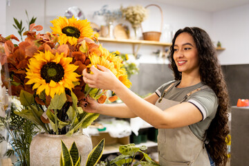 A young Latina woman works inside a pretty flower shop dressed in an apron.American woman is styling a sunflower plant.Concept of working woman. Women's Day.