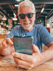 Wall Mural - Handsome smiling bearded senior man sitting at cafe table using mobile phone typing message or browsing internet