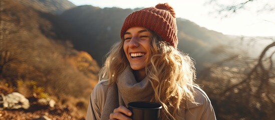 Wall Mural - A woman, wearing a cap and scarf, smiles happily while holding a cup of coffee, enjoying the scenic landscape. She looks relaxed and in a state of recreation and fun