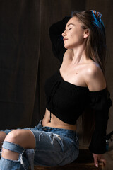 Classic studio portrait of a young brunette dressed in a black top, who is sitting on a chair against a black background.