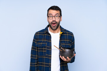 Wall Mural - Young caucasian man over blue background with surprise and shocked facial expression while holding a bowl of noodles with chopsticks