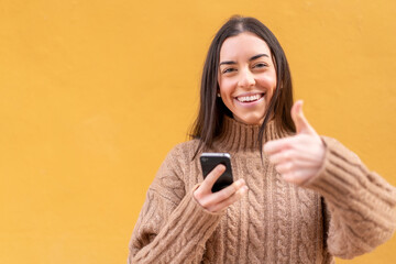 Wall Mural - Young brunette woman at outdoors using mobile phone while doing thumbs up