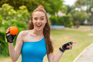 Poster - Young pretty sport girl holding an orange at outdoors surprised and pointing finger to the side