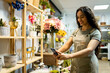 ©  Yistocking - A young Latina woman with long hair is working inside a beautiful flower shop dressed in an apron. The girl is taking a picture with her mobile phone to the products to upload it to the internet.