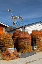 Multiple Stacks Of Industrial Crab Traps Or Pots For Commercial Fishing. The Round Nets Have Metal Frames With Orange Fishing Ropes. Long Metal Markers Are Stacked Behind The Traps Near The Red Shed.