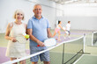 © JackF - Happy smiling elderly couple, man and woman in sportswear with rackets and balls in hands posing near net on indoor pickleball court after friendly match