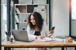 © Satori Studio - Businesswoman using her smartphone while reviewing documents, with a laptop open on the desk in a well-lit modern office.