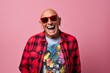 Handsome senior man in red sunglasses and a checkered shirt is laughing on a pink background.
