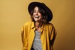 Cheerful young woman in hat laughing and looking at camera over yellow background