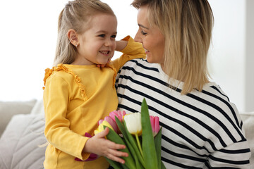 Wall Mural - Little daughter congratulating her mom with Mother`s Day at home. Woman holding bouquet of beautiful tulips