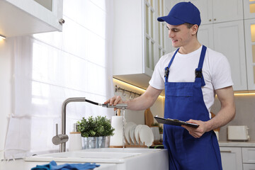 Canvas Print - Smiling plumber with clipboard examining faucet in kitchen