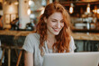 © Anek - A woman with red hair is sitting at a table with a laptop in front of her. She is smiling and she is enjoying herself