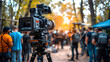 A high-end film camera set up on a tripod captures a scene on a movie set, surrounded by crew and autumn foliage