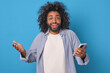 Young inspired positive Arabian man with phone happily spreads hands and looks at camera inviting you to download educational application to improve knowledge stands on blue studio background.