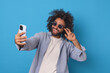 Young energetic cheerful Arabian man blogger in sunglasses records video blog on smartphone camera and greets subscribers urging them to subscribe to page on social network stands in blue studio