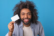 Young positive curly Arabian man with credit card in hands approaches camera and looks at you with smile urging use services of bank issuing loan posing on isolated blue background.