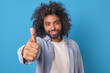 Young successful bearded Arabian man in casual clothes shows thumbs up appreciating your promotional product and motivating buyer to place order stands on isolated blue background. Student, millennial