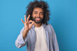 Young cheerful casual Arabian man with beard and mustache showing OK gesture with smile agreeing to plan you proposed or positively evaluating purchased product posing on isolated blue background.