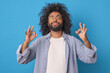 Young carefree smiling Arabian man with beard takes pose from yoga and zen practice to clear brain of harmful memories and gain positivity before work day stands on blue background.