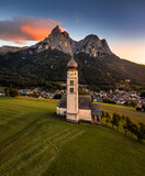 Fototapeta Londyn - Seis am Schlern, Italy - Aerial view of St. Valentin Church and famous Mount Sciliar mountain at background at sunset with golden and blue sky and warm sunlight at South Tyrol on a summer afternoon