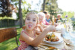 © Halfpoint - Portrait of a funny little girl sitting at table eating grilled food outdoors, sticking out tongue. Girl at family garden party.